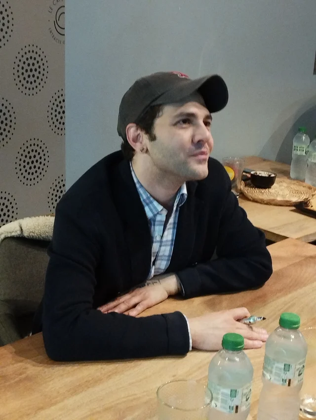 A man in a dark jacket and cap seated at a wooden table, looking away from the camera