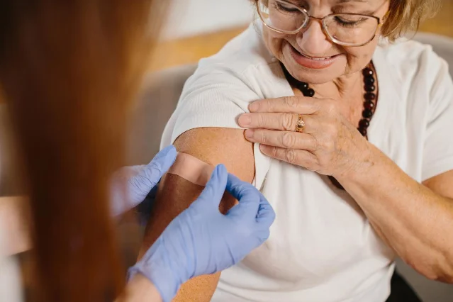 Older woman receiving a flu vaccine from a nurse, smiling as the nurse places a bandage on her upper arm