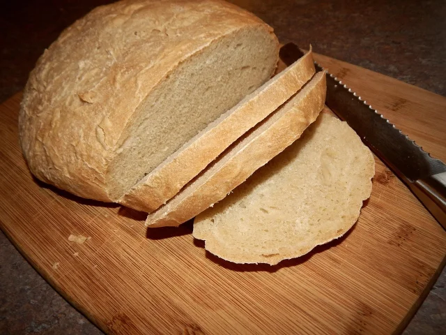 A round loaf cut into slices on a wooden board, with a serrated bread knife beside it