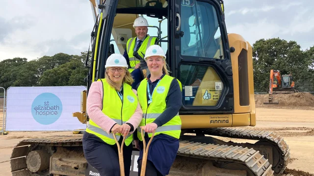 two women in hard hats and yellow high-visibility vests holding spades in front of a yellow excavator