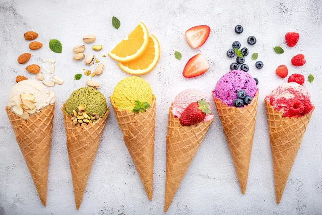 Six gelato cones in a flat-lay — the second from left is a green pistachio scoop with whole pistachios and almonds scattered around it