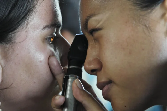 A clinician using an ophthalmoscope to examine a patient's eye at close range, retinal light visible in the patient's pupil