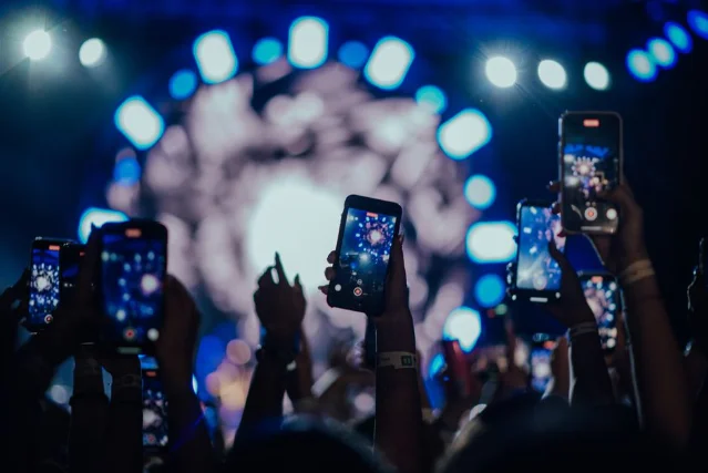 Crowd raising phones to film a concert, stage lights blazing blue in the background