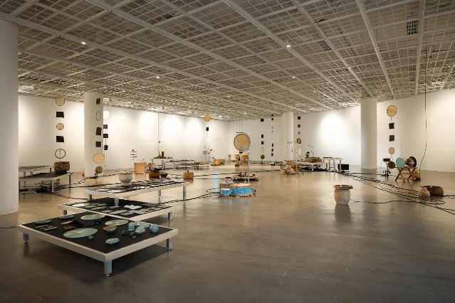 一间白色展厅里摆满大小鼓、铜碗、瓷盘与悬挂的圆盘,地面上电线连成一片