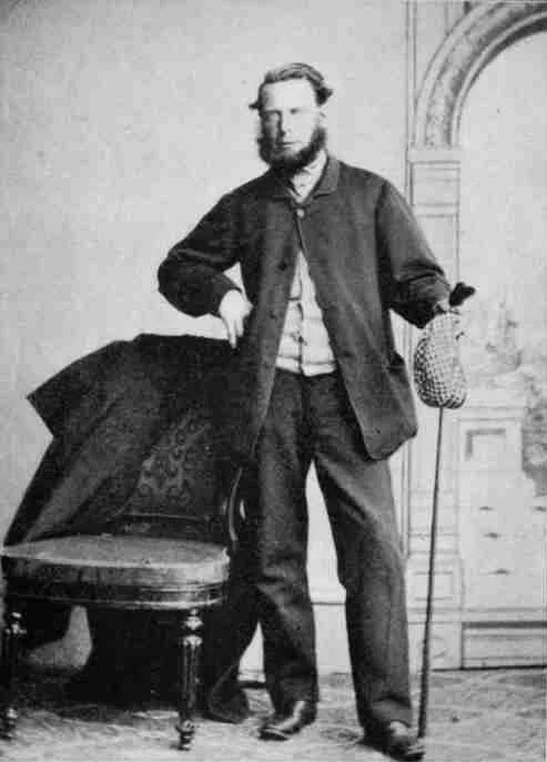 Tom Morris, bearded and in a dark jacket, standing beside a draped chair with a club