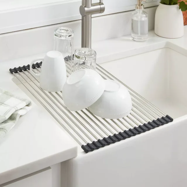A roll-away dish drying rack of stainless steel rods with black silicone ends spanning a white kitchen sink, holding white bowls, a mug, and glasses.