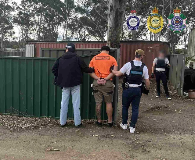 Three AFP and Queensland Police officers restraining a handcuffed man in an orange shirt against a green fence line of shipping containers in a scrubby bush setting, AFP and QPS badge overlays in the top-right corner