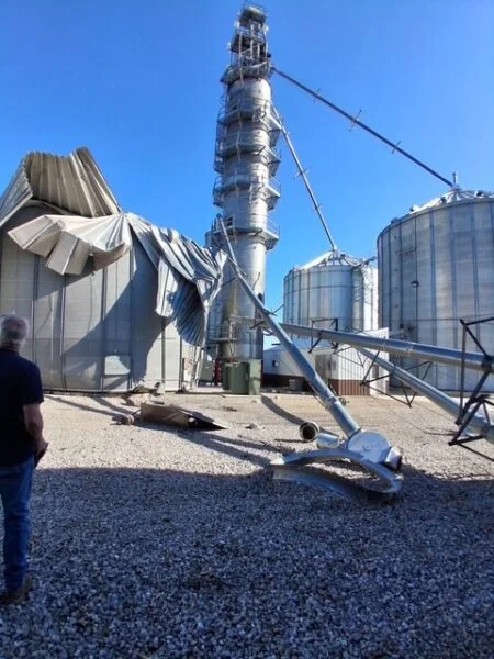 Storm-damaged grain bins at a Midwest facility, with structural panels blown off and auger equipment displaced