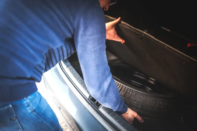A person in a blue sweater holding up the load floor of a car's trunk, revealing a black steel spare wheel resting in the well beneath