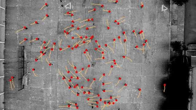 Overhead drone footage of a concrete plaza with dozens of pedestrians shown as red dots, each trailing a tan trajectory line that curves predominantly counterclockwise