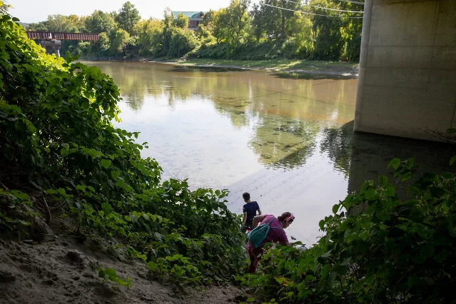 Two people working down a steep, overgrown bank to the waterline of a broad river, with a road bridge overhead at right and a single-storey building on the far bank