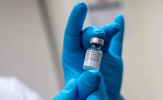 A pair of gloved hands holding a small glass vaccine vial