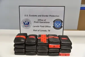 Twenty dark rectangular cocaine bricks wrapped in black plastic with orange tape bands, stacked in three rows on a table, with a CBP "Laredo Field Office / Port of Laredo, TX" sign in the background