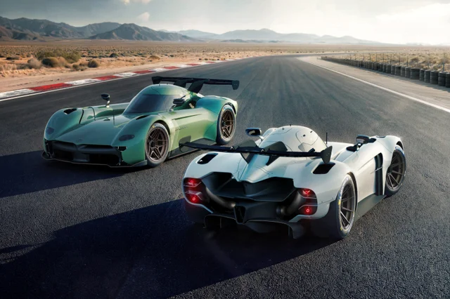 Two low, open-cockpit sports cars, one green and one white, parked on a desert race track with mountains behind