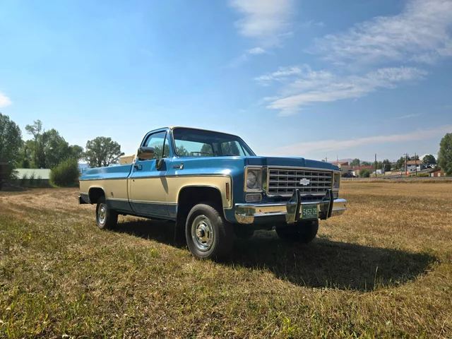 A two-tone blue and cream 1976 Chevrolet K10 pickup parked in a dry grass field under a blue sky