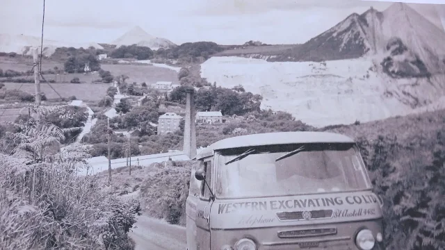 A black-and-white photograph of houses and trees in a valley, with white clay tips on the hills behind and a van on the lane in the foreground