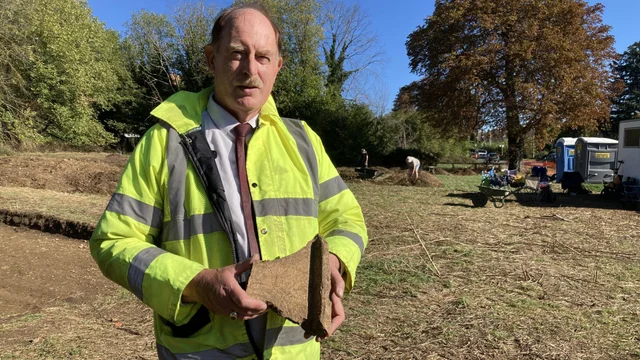 a man in a high-visibility jacket holding a piece of brown tile in a field, with a wheelbarrow and portable toilets behind him
