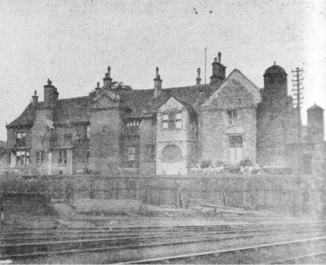A black-and-white photograph of a large stone manor house with gabled roofs, tall chimneys and a domed turret, standing behind a fence and a set of railway tracks