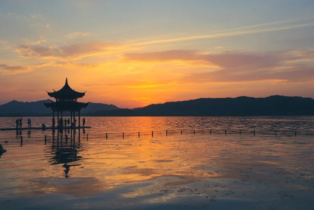A traditional pavilion silhouetted over West Lake in Hangzhou at sunset, mountains visible in the distance