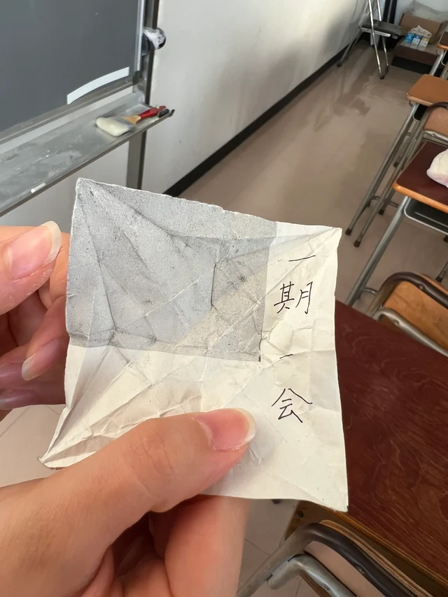 Two hands holding up a crumpled square of white paper with four black characters written in a column down its right side