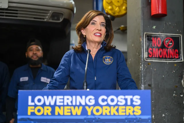 New York Governor Hochul speaking at an auto repair shop, with a banner reading "LOWERING COSTS FOR NEW YORKERS"