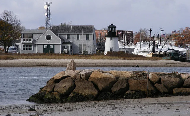 Rocky stone jetty in the foreground, New England harbor with small lighthouse, gray-shingled houses and boats behind