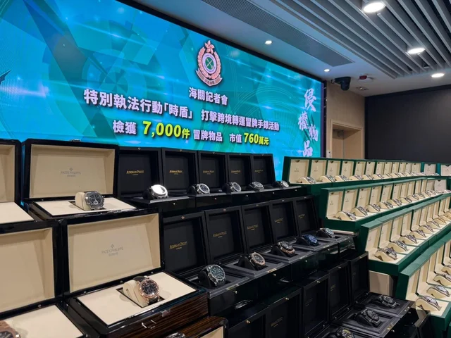 Rows of open watch boxes holding suspected counterfeit watches, with a Hong Kong Customs operation screen behind them showing 7,000 items and HK$7.6 million
