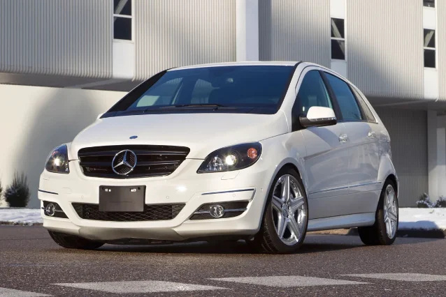 A white Mercedes-Benz B-Class hatchback photographed three-quarter front on asphalt with a corrugated metal building behind