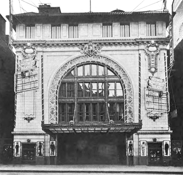 A black-and-white photograph of a theatre facade with a tall glass arch above a canopy, and vertical signs reading Eltinge 42nd Street Theatre