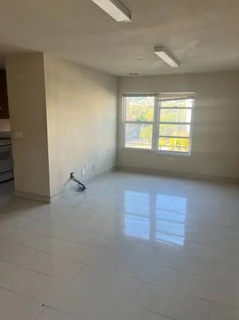 10879 San Pablo Ave #114 interior — empty room with glossy tile floor, corner window showing greenery, recessed ceiling lights, partial kitchen view