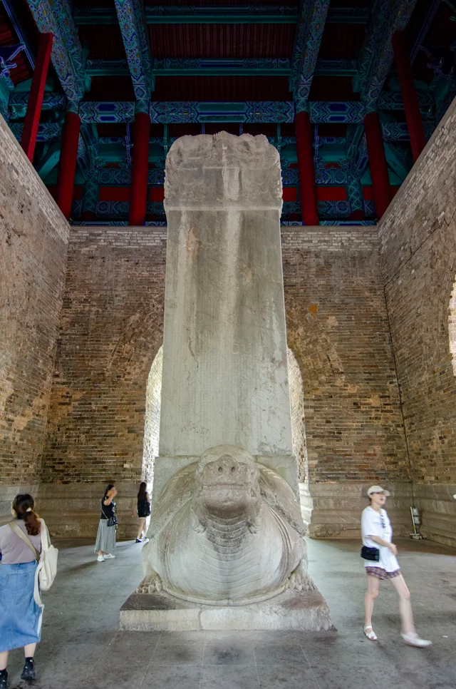 A very large stone stele standing on the back of a carved stone tortoise inside a brick hall, with visitors for scale