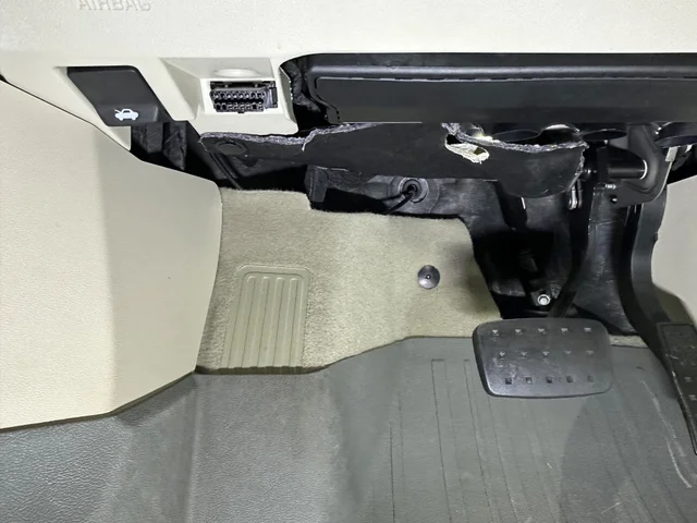 Looking up into the driver's side footwell of a car with light beige trim, the rectangular 16-pin diagnostic socket set into the panel beneath the dashboard, floor mat and pedals below