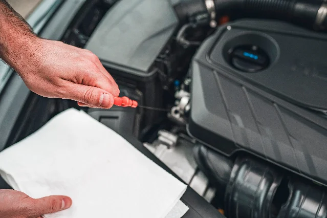 A hand holding a red-handled oil dipstick it has just drawn out of the engine, above a white paper towel, with the engine bay behind