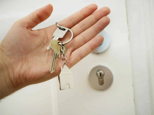 A hand holding two keys on a ring with a house-shaped keyring, in front of a closed white door.