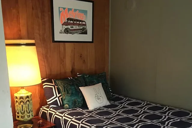 A bed with navy-and-white patterned bedding and teal cushions, under a framed Melvins poster on a wood-panelled wall, beside a lit table lamp