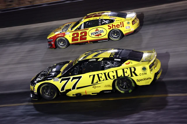 Two NASCAR Cup cars racing side by side under the lights, the No. 22 Ford on the inside of the No. 77 Chevrolet