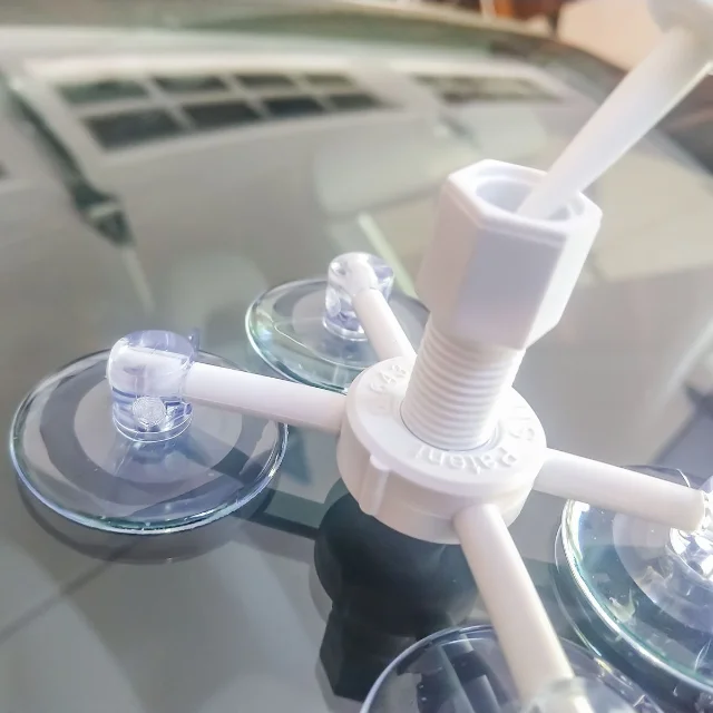 A white plastic resin chamber with a threaded body and a square plunger knob attached at the centre of the four-cup fixture, sitting on a windshield in daylight