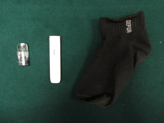 A clear etomidate capsule and a slim white pod-style e-cigarette device laid out on a green inspection surface, with a black ankle sock alongside them