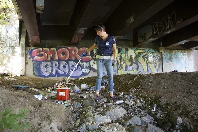 A woman in a T-shirt and jeans stands on a rocky bank under a road bridge, using a litter picker to lift debris into a red bucket, with graffiti on the concrete wall behind her