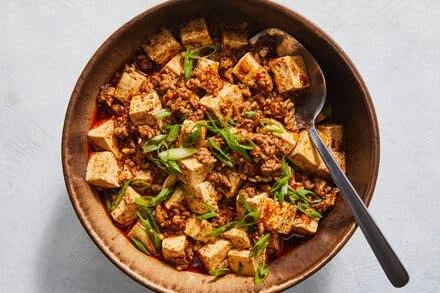 Bowen Yang's Mapo Tofu in a copper-toned bowl with firm tofu cubes, ground pork in red sauce, and sliced scallions on top