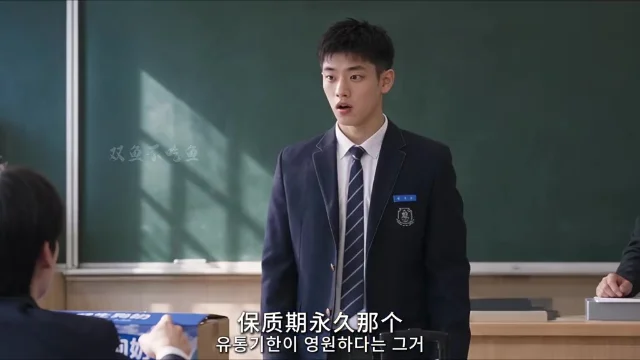 教室里,一名身穿深色校服西装的男生站在绿色黑板前,手中拿着一个小纸盒,画面下方是中韩双语字幕。