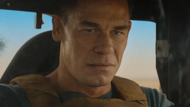 John Cena in a tan vest seated inside a vehicle, looking out through the window