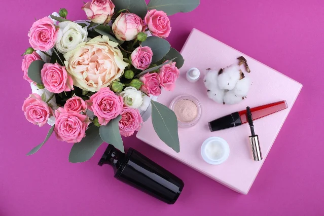 Beauty skincare flatlay with pink roses, cream jars, mascara, and a dark serum bottle on a pink background