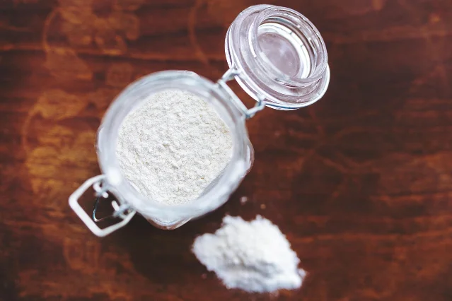 An open glass clip-top jar of flour standing on a dark wooden surface