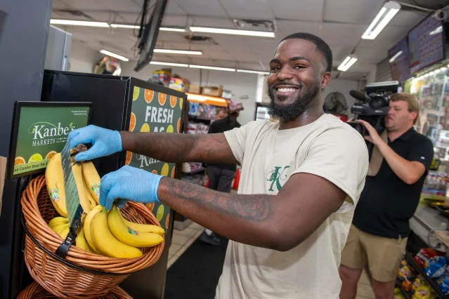 A worker in gloves arranging bananas in a wicker basket beside a cooler at a corner store