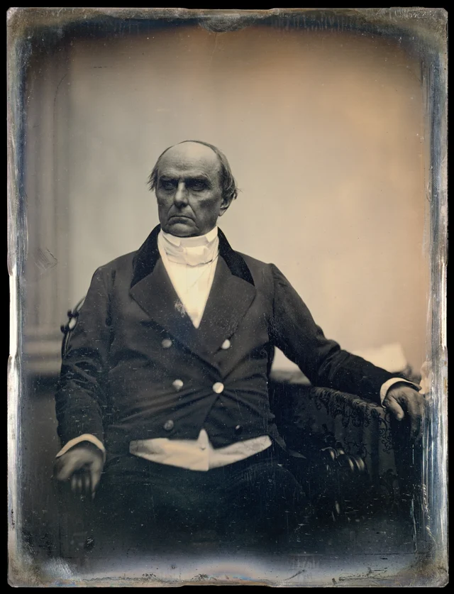 A seated man in a dark double-breasted coat and a high white collar, facing the camera