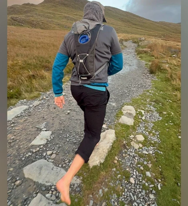 a barefoot walker in a grey hoodie and rolled-up trousers, walking away up a stony mountain path