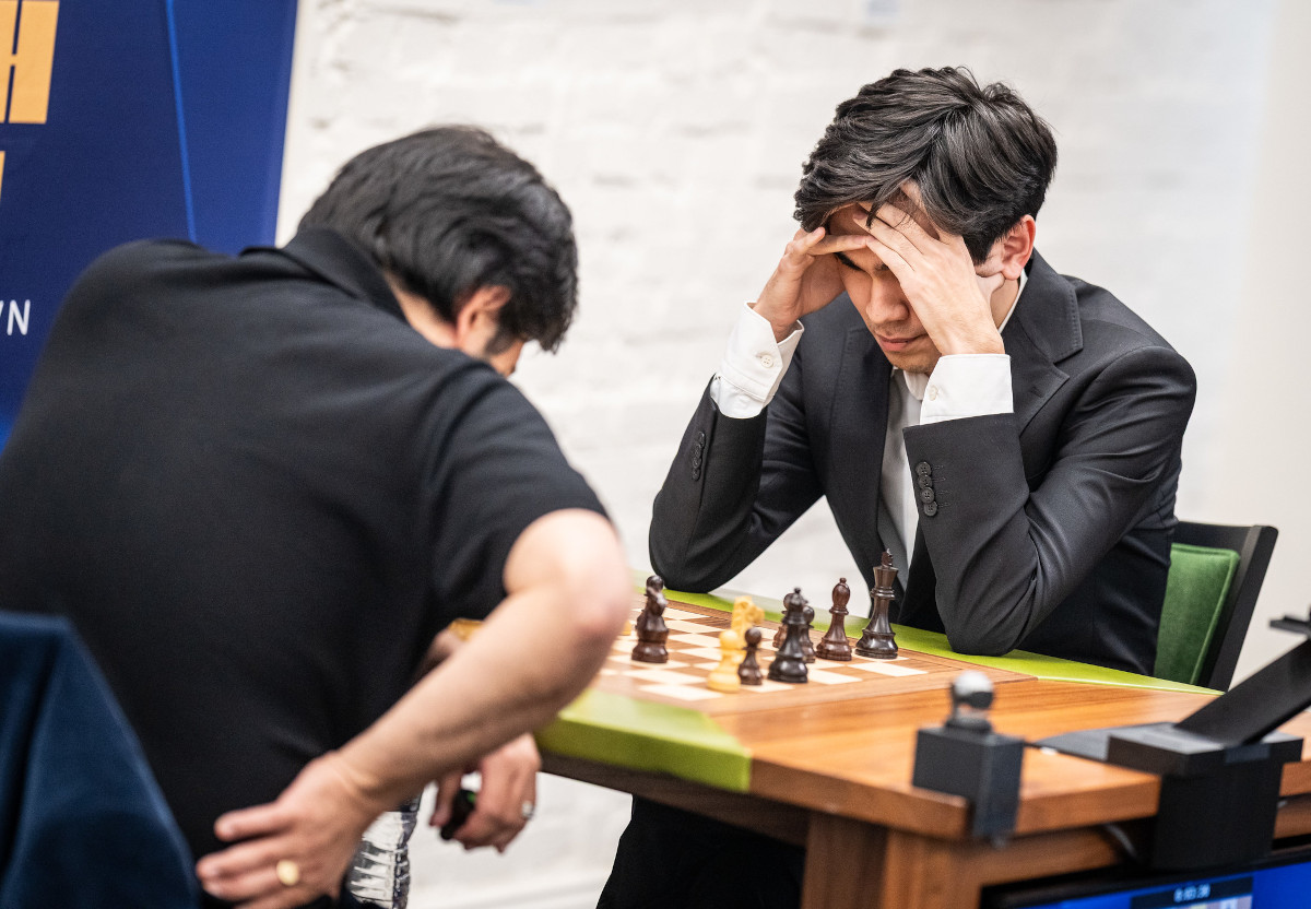 Two players at a chessboard in a bright room, the nearer player leaning forward with his head in his hands
