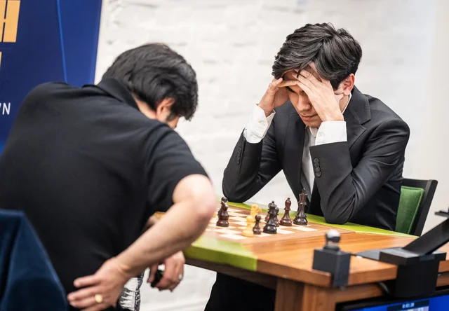 Two players at a chessboard in a bright room, the nearer player leaning forward with his head in his hands