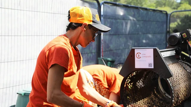 A Criw Compostio worker feeds material into a branded processing machine while another worker sorts the pile behind them.